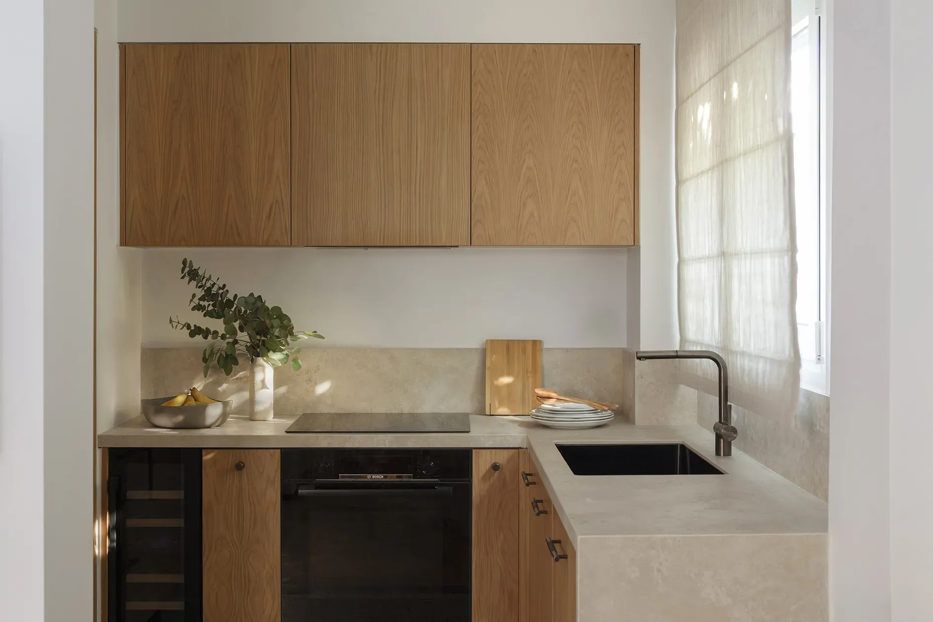 Oak kitchen cabinetry with stone worktops and daylight through a softly screened window