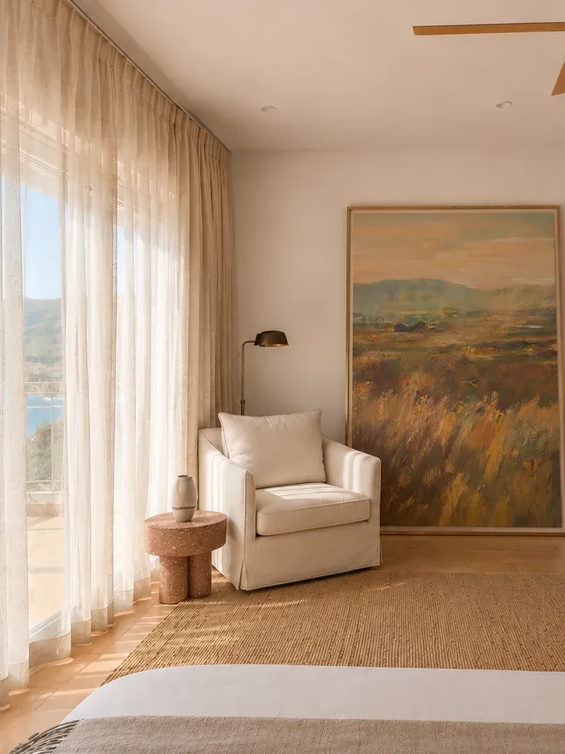 Sunlit bedroom corner with a linen armchair, landscape artwork and views across Limnos